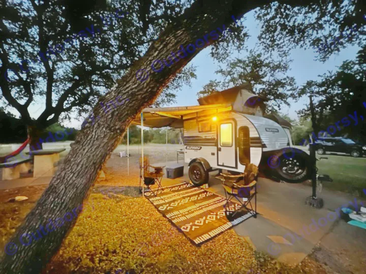 Cozy night camping with an RV rental. A lit Sunray travel trailer, awning, rug, & chairs under trees.. Sunset Park RV SunRay 2023