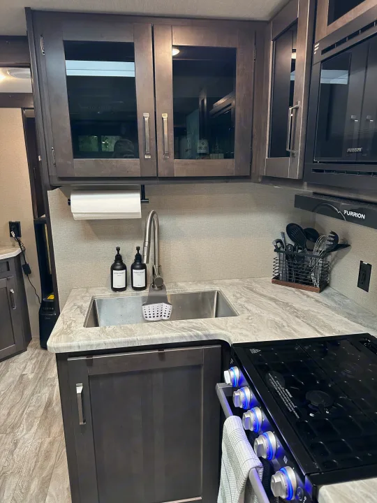Compact rv rental kitchen with dark wood cabinets, sink, and modern cooktop. Essential for convenient mobile living.. Grand Design Transcend Xplor 2022