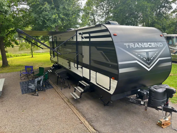 This RV rental, a Transcend travel trailer, is set up at a scenic lakeside campsite with an extended awning and chairs.. Grand Design Transcend Xplor 2022