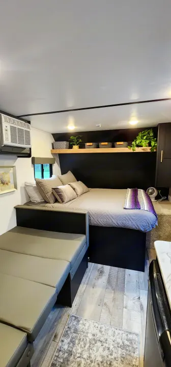 Inside an RV rental, a compact bedroom/living area. Features a bed with pillows, a grey bench, wooden floor, and a shelf with plants.. Keystone RV Springdale 2022
