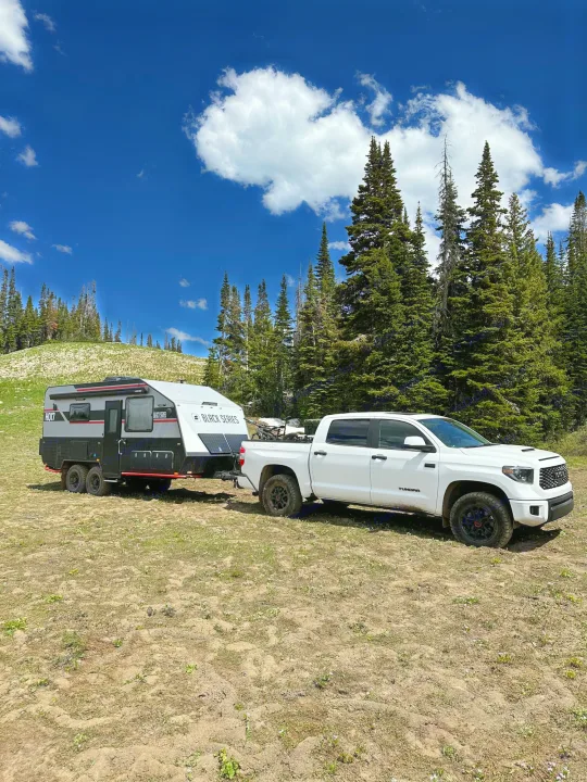 White pickup truck with a Black Series camper, ideal for an RV rental trip, parked in a sunny forest.. Black Series Black Series Trailer 2022