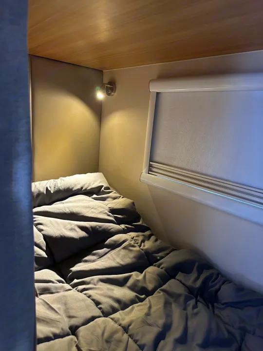 Cozy RV rental bunk bed with a warm reading light, gray comforter, and window blind in a compact sleeping area.. Black Series Black Series Trailer 2022