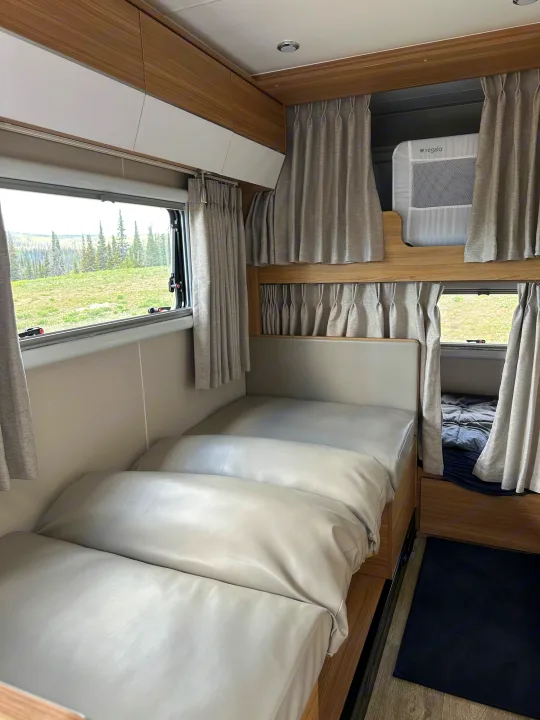 Inside an **rv rental**: two beds, upper bunk with a "Regalo" baby cot, curtains, storage, and a scenic forest view outside.. Black Series Black Series Trailer 2022