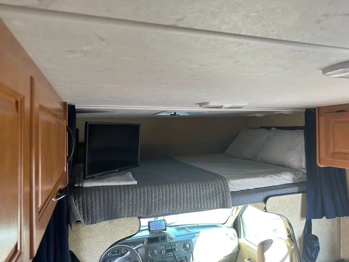 Inside an RV rental: a cozy cab-over bunk bed with a TV, pillows, and quilt, offering a comfy sleeping space above the driver's cabin.. Thor Majestic 2015
