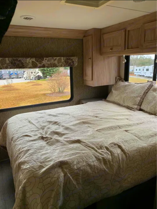 An RV rental's bedroom, featuring a comfy bed with patterned linens, overhead cabinets, and a window view of an RV park.. Forester LE 2018