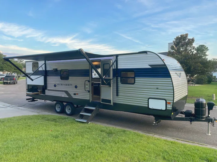 Grey and blue travel trailer, great for an RV rental, parked roadside with awning extended & steps down.. Forest River Prime time Avenger 2023