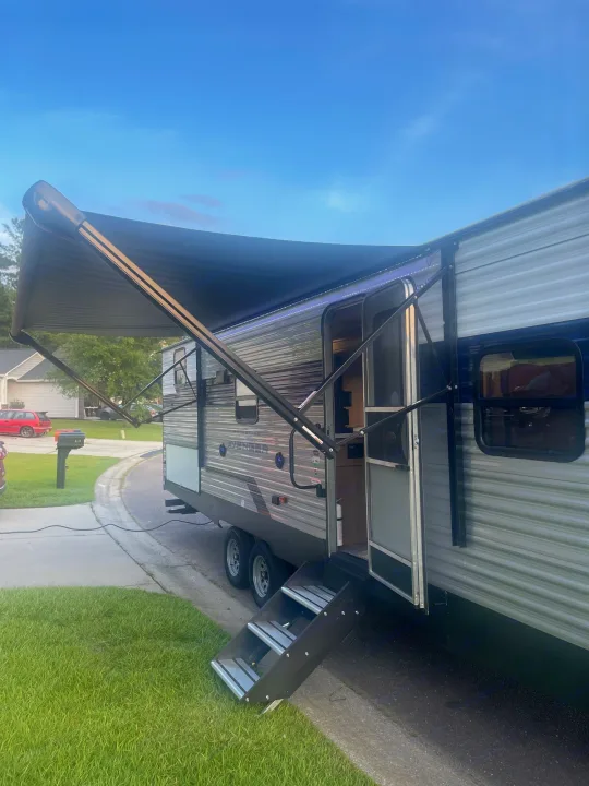 Travel trailer RV rental with an extended awning and deployed steps, parked on a driveway in a sunny residential area.. Forest River Prime time Avenger 2023