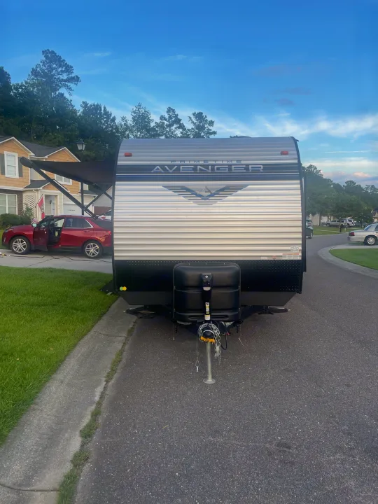 A Prime Time Avenger RV rental is parked on a suburban street, with a red car and houses visible under a blue sky.. Forest River Prime time Avenger 2023