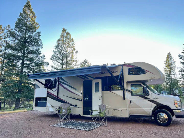 RV rental parked at a serene campsite with awning extended. Two chairs and a patterned rug are set out on the gravel, surrounded by tall pine trees.. Thor Four Winds 2020