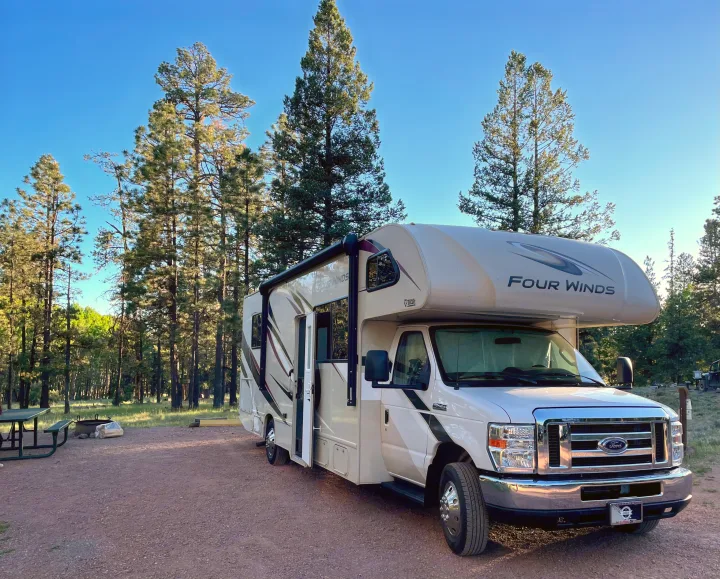 An RV rental is parked at a scenic campsite surrounded by tall pine trees under a clear blue sky, with a picnic table and fire pit.. Thor Four Winds 2020