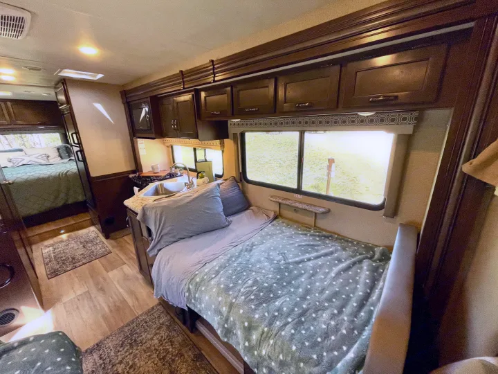 Cozy RV rental interior features a bed with green blanket, compact kitchen with sink, overhead cabinets, and wood flooring.. Thor Four Winds 2020