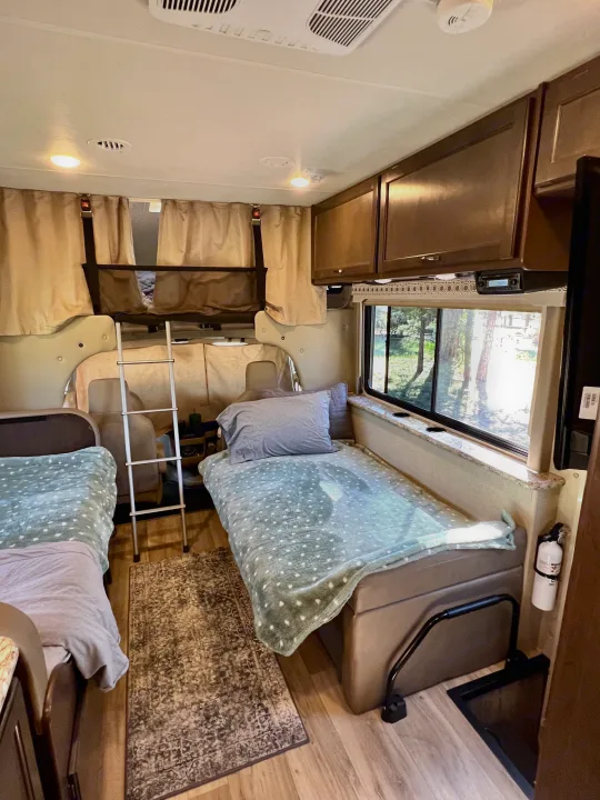Cozy rv rental interior with bunk beds, ladder, window view of trees, and a fire extinguisher, ready for travel.. Thor Four Winds 2020