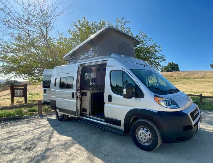 Silver pop-top RV rental, its side door open showing the interior, parked on a sunny dirt road.. Winnebago Solis 2021
