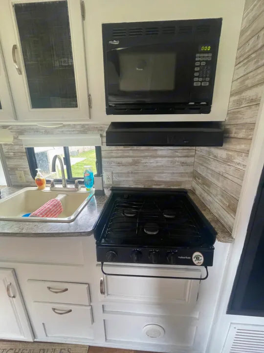 Modern RV rental kitchen: white cabinets, light wood backsplash, High Pointe microwave, gas stove, and sink area.. Forest River Wildwood X-Lite 2016