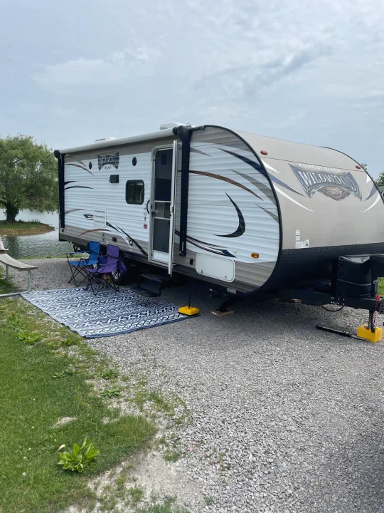 This inviting RV rental campsite by the water features a travel trailer, outdoor mat, and chairs, ready for relaxation.. Forest River Wildwood X-Lite 2016