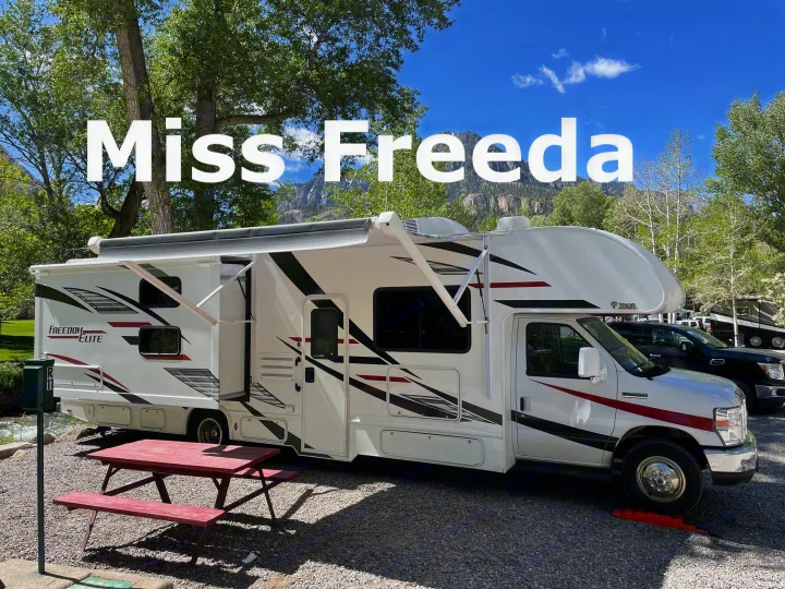 Miss Freeda, a white RV rental, parked by a river with an extended awning and red picnic table, under sunny skies.. Thor Freedom Elite 2020