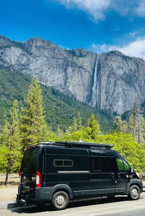 Black RV rental parked near majestic Yosemite Falls and granite mountains, surrounded by lush green trees under a blue sky.. Dodge Ram Van Roadtrek 2019