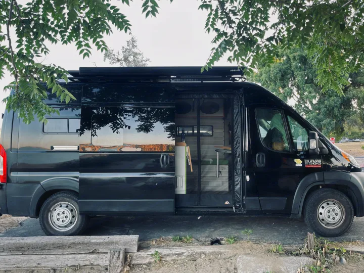 Black Dodge Ram camper van for RV rental, with side door open revealing its custom interior. Parked under trees.. Dodge Ram Van Roadtrek 2019
