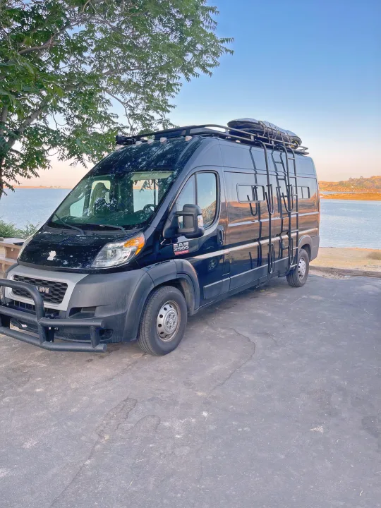 Black Ram ProMaster 2500 RV rental parked lakeside, equipped with a roof rack & bull bar, perfect for outdoor travel.. Dodge Ram Van Roadtrek 2019