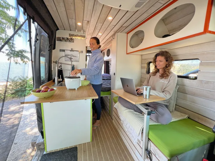 Two women in an RV rental: one cooking at the kitchen, the other working on a laptop in a cozy, bright camper van.. Dodge Ram Van Roadtrek 2019