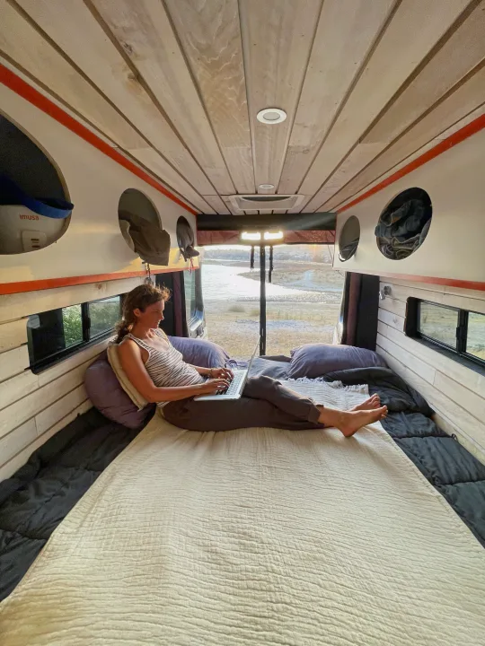 Working remotely from a cozy RV rental: A woman uses her laptop on a bed inside a camper van, overlooking a scenic landscape.. Dodge Ram Van Roadtrek 2019