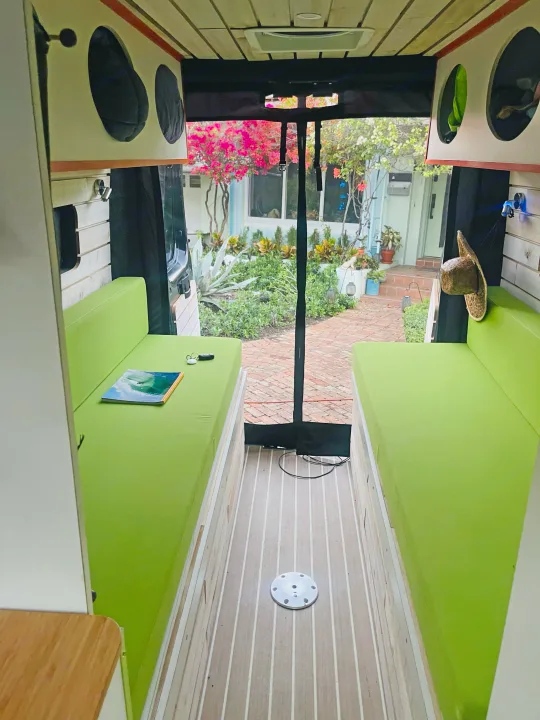 Vibrant green benches and wood-plank floor inside an RV rental, with a clear view of a beautiful outdoor garden.. Dodge Ram Van Roadtrek 2019