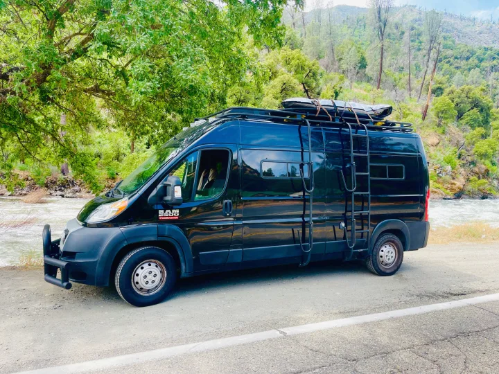 Black RV rental van beside a vibrant river, surrounded by lush mountains and trees. Equipped for adventure.. Dodge Ram Van Roadtrek 2019