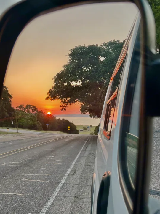 RV rental side mirror reflects a fiery sunset over a lake, with a road and trees. The orange sun hangs low above the horizon.. Dodge RAM Promaster 2017