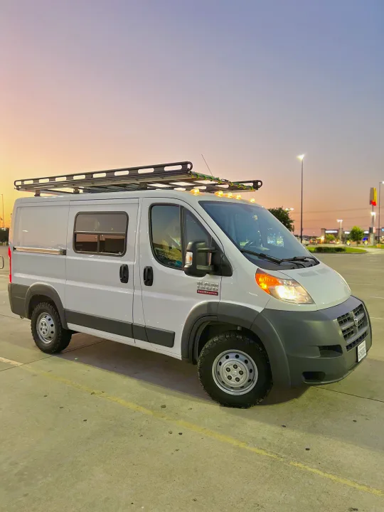 A white Ram ProMaster camper van with a roof rack and rugged tires, parked in a lot at dusk. Perfect for an rv rental.. Dodge RAM Promaster 2017