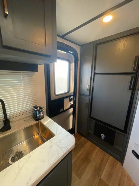 Modern RV rental kitchen featuring a stainless steel sink, marble-patterned counter, dark gray cabinets, and an integrated refrigerator.. Keystone RV Bullet Crossfire 2022