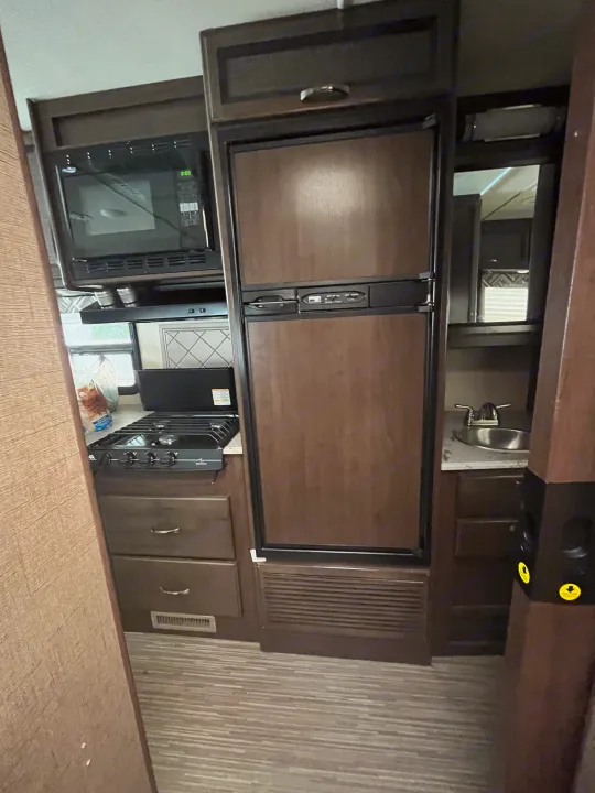Inside this RV rental: compact kitchen featuring a microwave, stove, fridge, and small sink. Wood-tone cabinets.. Thor Axis 2017