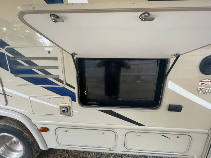 Side of an RV rental with an open outdoor TV compartment, storage, and utility vents visible on the beige exterior.. Thor Axis 2017