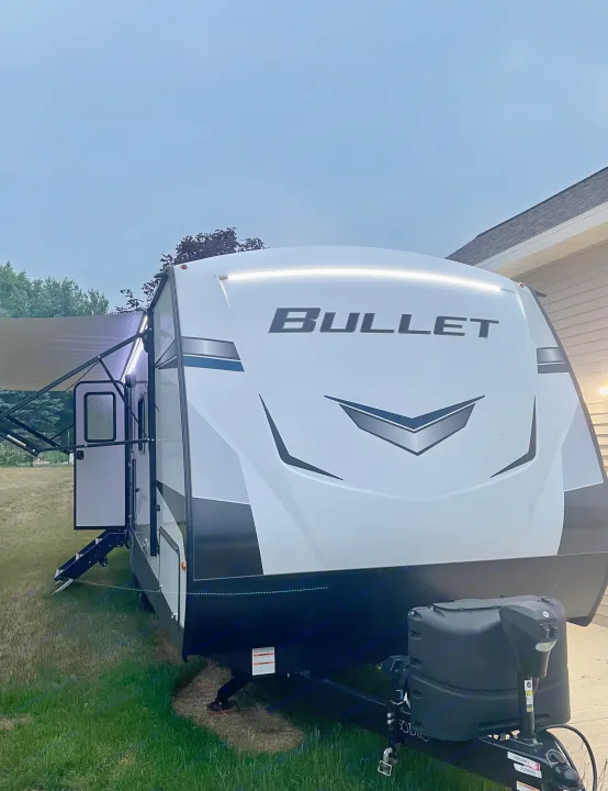 A bright white Bullet travel trailer, great as an RV rental, seen at dusk with its awning extended and lights illuminated.. Keystone RV Bullet Ultra Lite 2022