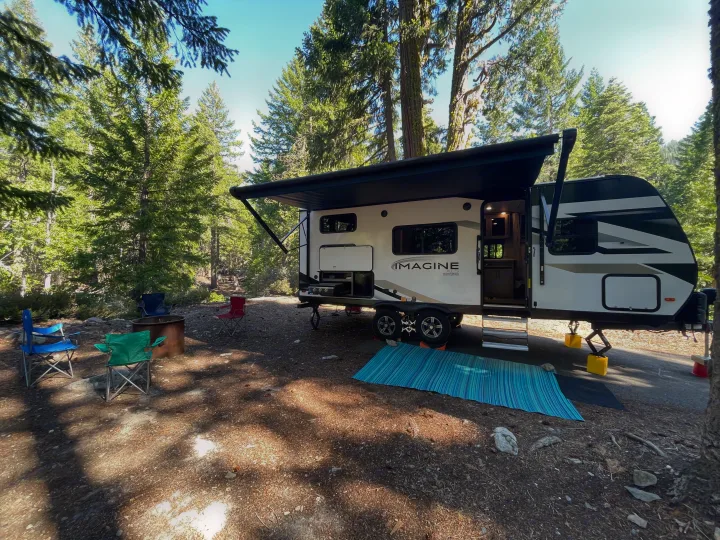 RV rental travel trailer parked at a serene forest campsite with an extended awning, outdoor kitchen, fire pit, and chairs.. Grand Design Imagine XLS 2023