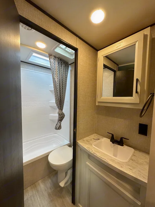 View of a modern RV rental bathroom, featuring a white tub/shower with a patterned curtain, toilet, and vanity with sink & mirror.. Jayco Jay Flight SLX 2022