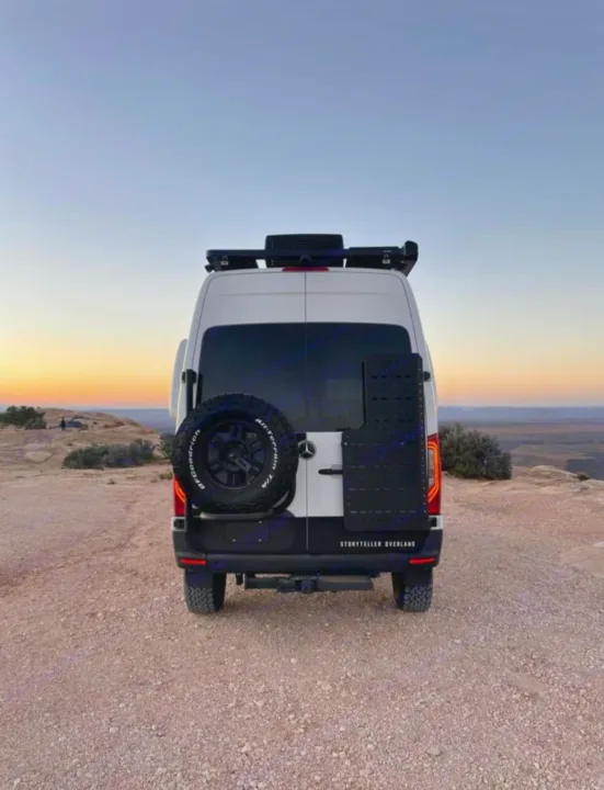 Rear view of a white Storyteller Overland adventure van (RV rental) with spare tire, parked on a dusty desert overlook at sunset.. Mercedes-Benz sprinter 2023