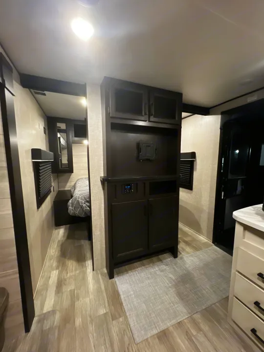 Modern RV rental interior with dark built-in cabinetry, an entertainment system, and TV mount. Light wood flooring and kitchen area visible.. Jayco Jay Flight 2023