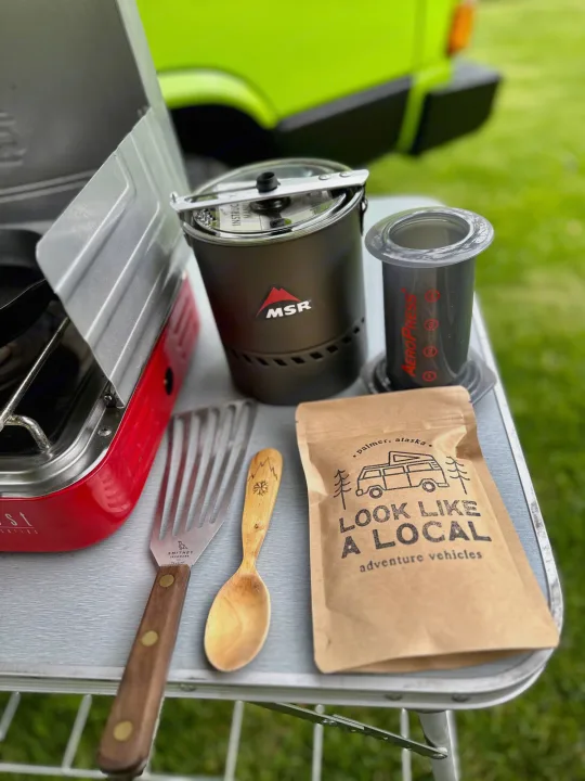 RV rental essentials: MSR camp kitchen gear, AeroPress, and local coffee on a table, with a green adventure vehicle in the background.. Volkswagen Westfalia 1984