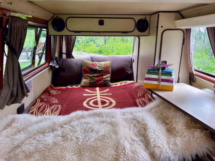 RV rental interior featuring a cozy bed with a furry blanket, a stack of books, and a beautiful green nature view.. Volkswagen Westfalia 1984