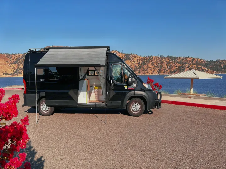 Black custom camper van with extended awning, parked by a scenic lake. Perfect RV rental for outdoor adventures!. Dodge Ram Van Roadtrek 2019