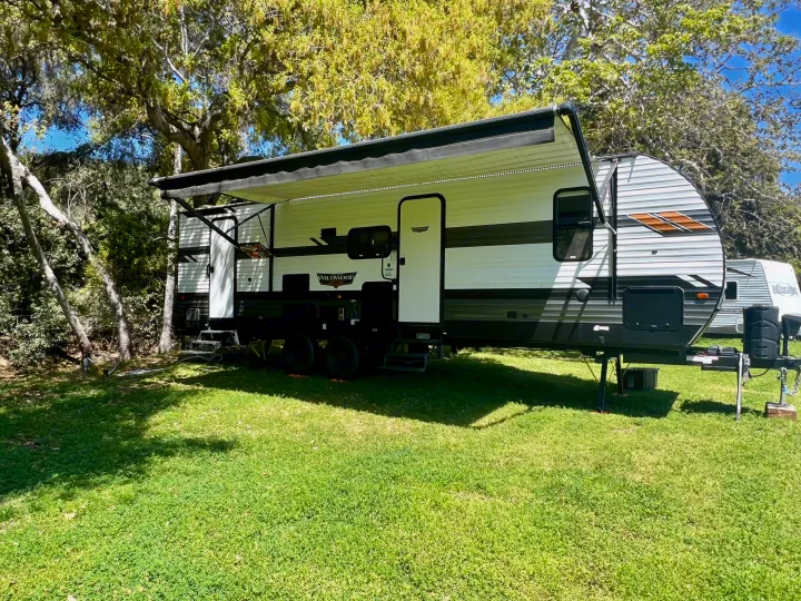 This Wildwood RV rental is set up with its awning extended on bright green grass amidst trees, ready for camping.. Forest River Wildwood 2021
