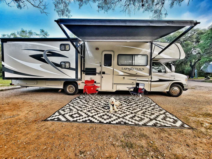 RV rental set up at a scenic campsite. An extended awning shades a mat with chairs and a fluffy white dog.. Coachmen Leprechaun 320BH 2014