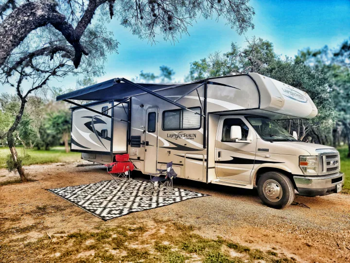 An RV rental, a Coachmen Leprechaun, is set up at a scenic campsite with chairs and a mat, perfect for outdoor relaxation.. Coachmen Leprechaun 320BH 2014
