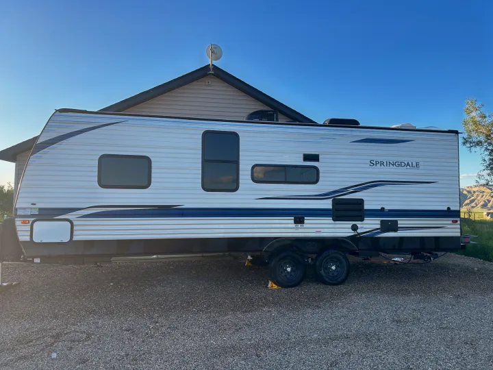 A white Springdale travel trailer, ideal for RV rental adventures, is parked on a gravel lot under a clear sky. A perfect getaway vehicle.. Keystone RV Springdale 2021