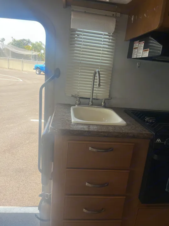 RV rental kitchen: sink, faucet, stove, countertop, and drawers. Paper towels above window, outdoor view with blue truck.. Thor Four Winds 2015