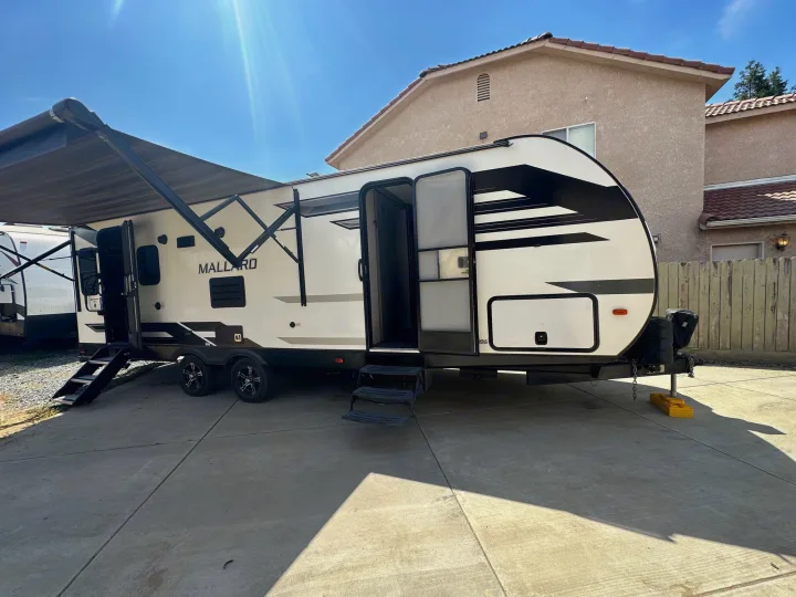 This white & black Mallard RV rental features an extended awning & open door, parked on a sunny driveway, ready for travel.. Heartland Other 2021