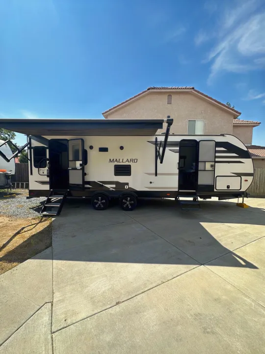 A white and black Mallard RV rental, with its awning extended and entry doors open, parked on a concrete driveway.. Heartland Other 2021