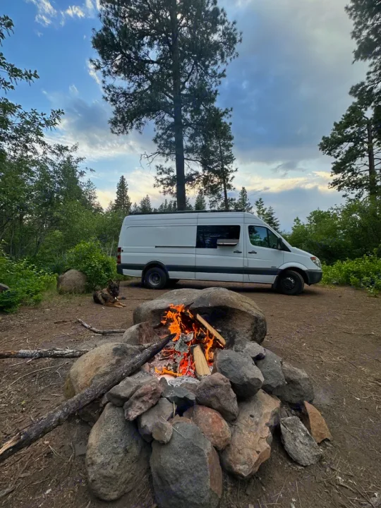 Cozy campsite evening with an RV rental, featuring a white camper van, a crackling campfire, and a resting dog.. Mercedes Sprinter 2010