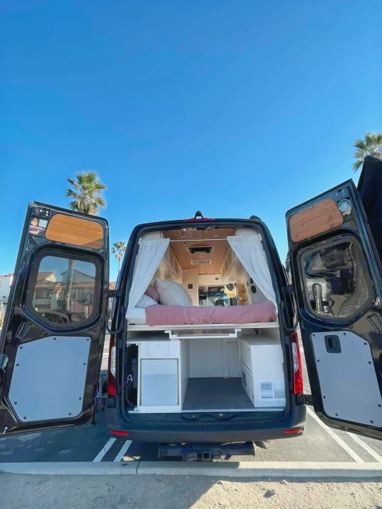 This rv rental camper van showcases a cozy bed & ample storage through its open rear doors, ready for a road trip.. Mercedes-Benz Sprinter 2021