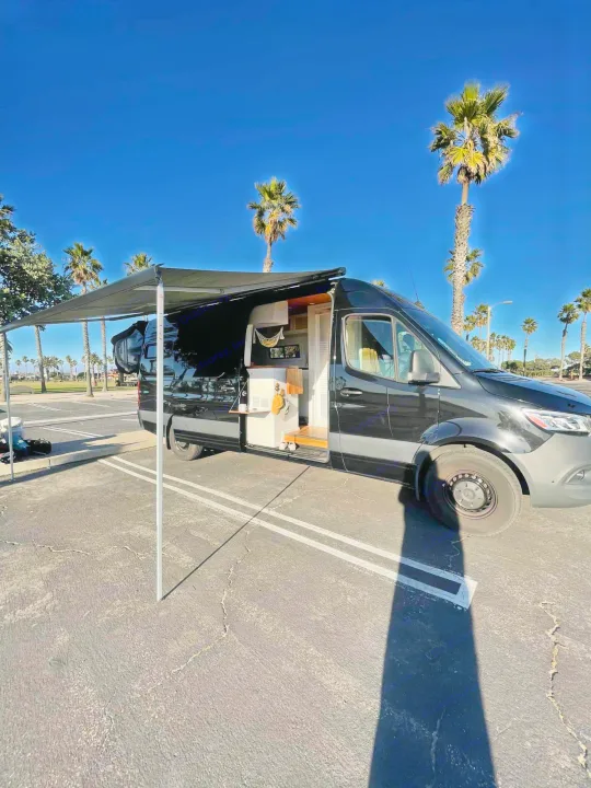 Black RV rental van with extended awning. Interior visible as it's parked by palm trees on a sunny day.. Mercedes-Benz Sprinter 2021